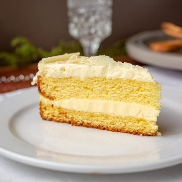 A slice of vanilla cake with white frosting and white chocolate shavings on a white plate.