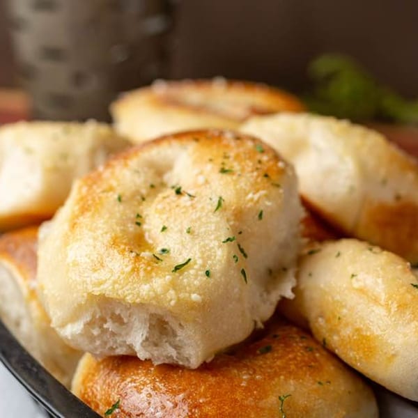 Close-up of golden-brown garlic knots, sprinkled with parmesan cheese and parsley, in a black basket.