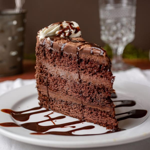 A decadent slice of chocolate cake with chocolate frosting, whipped cream, and chocolate drizzle.